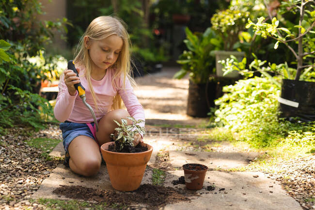 Ein kaukasisches Mädchen mit langen blonden Haaren genießt die Zeit in einem sonnigen Garten, erkundet, pflanzt einen Sämling in einen Topf mit Gartengabel — Stockfoto