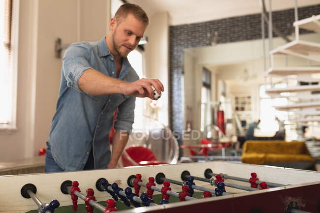 Vorderansicht eines jungen kaukasischen Mannes, der es sich zu Hause gemütlich macht, in seinem Wohnzimmer steht und Tischfußball spielt. — Stockfoto