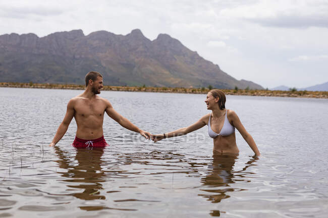 Передній вид на кавказьку пару добре проводить час у подорожі в гори, стоячи в озері, тримаючись за руки, дивлячись один на одного. — стокове фото
