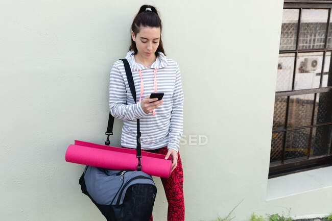 Front view of a fit Caucasian woman on her way to fitness training, carrying a sports bag and a yoga mat, standing on a street against a wall, using her smartphone — Stock Photo