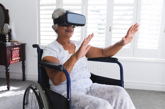 A senior retired African American woman at home, sitting in a wheelchair in her nightclothes in front of a window on a sunny day using a VR headset with her arms outstretched in front of her, self isolating during coronavirus covid19 pandemic — Stock Photo