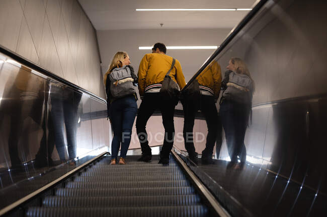 Vista trasera de ángulo bajo de una pareja caucásica en la ciudad, subiendo en la estación de metro con una escalera mecánica. - foto de stock