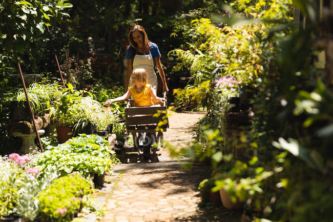 A Caucasian woman wearing an apron and her daughter enjoying time in a sunny garden, the daughter sitting in a wheelbarrow while the mother pushes her along — Stock Photo