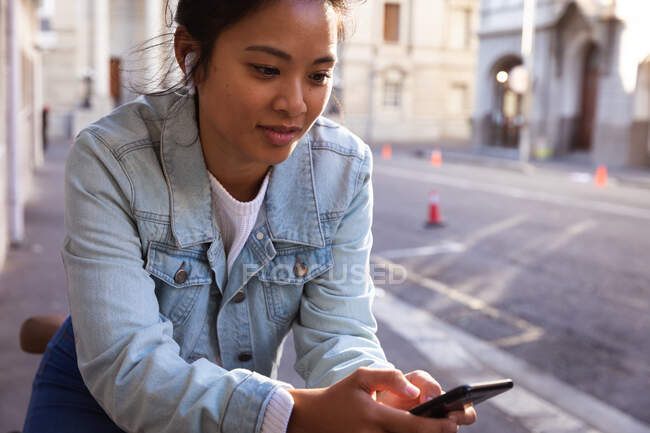 Front view close up of a mixed race woman with long dark hair out and about in the city streets during the day, leaning on her bicycle and using a smartphone with earphone on with buildings in the background. — стоковое фото
