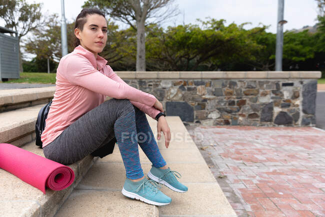 Бічний вид на підходящу кавказьку жінку на шляху до тренування фітнесу в похмурий день, зі спортивним пакетом і кішкою йоги поруч з нею, сидячи на сходах у парку. — стокове фото