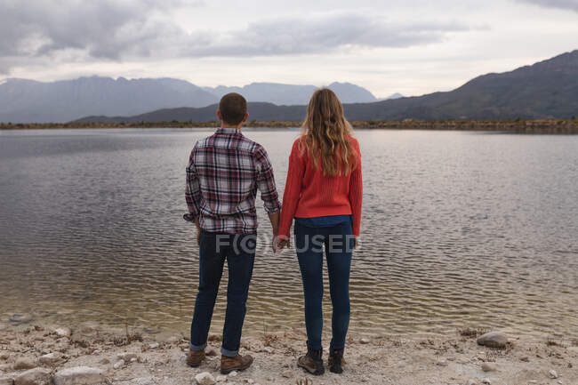 Vista posteriore da vicino di una coppia caucasica che si diverte durante un viaggio in montagna, in piedi su una riva del lago, tenendosi per mano — Foto stock