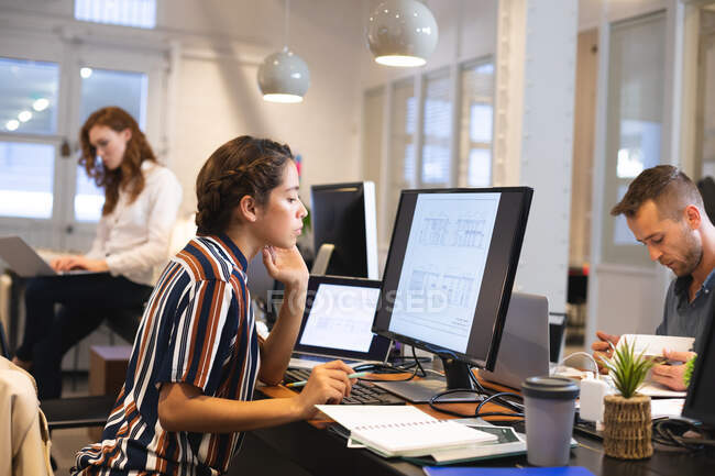 Творческая женщина смешанной расы работает в обычном современном офисе, сидит за столом и пользуется компьютером с коллегами, работающими на заднем плане — стоковое фото