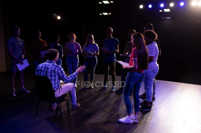 Бічний погляд на кавказького вчителя середньої школи, який сидить на стільці на сцені в порожньому шкільному театрі під час репетицій для виступу, готуючи багатоетнічну групу студентів-підлітків для виступу. — стокове фото
