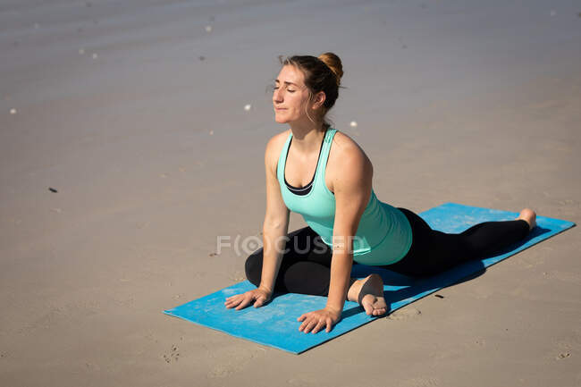Vista lateral de una atractiva mujer caucásica, vestida con ropa deportiva, practicando yoga sobre esterilla de yoga, estirándose en posición de yoga, en la playa soleada. - foto de stock