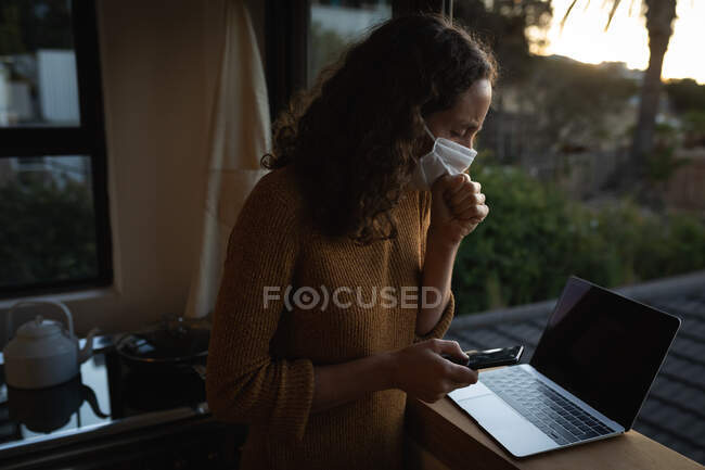 Белая женщина проводит время дома, изолируя себя, надевая маску для лица, сидя у окна и работая с ноутбуком и смартфоном, прикрывая рот во время кашля. — стоковое фото