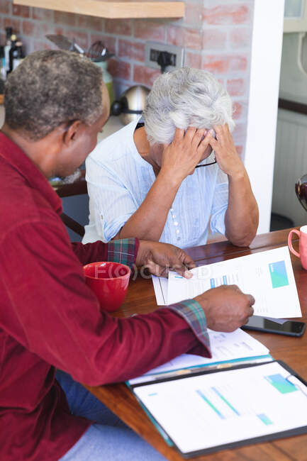 Una pareja de ancianos afroamericanos jubilados sentados en una mesa en su comedor bebiendo café, mirando el papeleo y discutiendo sus finanzas, la mujer con la cabeza en las manos, en casa juntos aislándose durante la pandemia del coronavirus covid19 - foto de stock