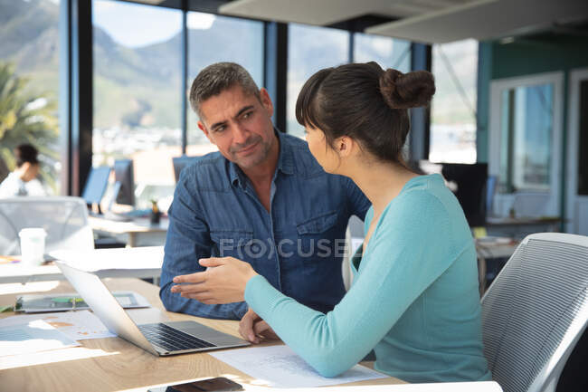 An Asian businesswoman and a Caucasian businessman working in a modern office, using a laptop computer and talking, with their colleagues working in the background — Stock Photo