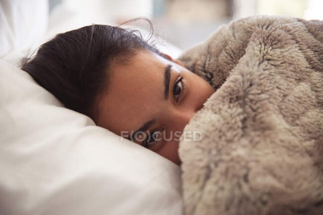 Mixed race woman spending time at home self isolating and social distancing in quarantine lockdown during coronavirus covid 19 epidemic, lying in bed on pillow covered with soft blanket in bedroom. — Stock Photo