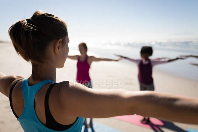 Vista trasera de la mujer caucásica, vistiendo ropa deportiva, practicando yoga con los brazos extendidos en la playa soleada con sus amigos estirándose en el fondo. - foto de stock