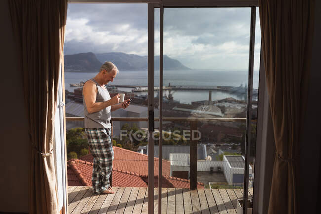 Vista laterale di un anziano caucasico che si rilassa a casa indossando un gilet e pantaloni pigiama, in piedi sul balcone con un telefono cellulare e bere caffè — Foto stock