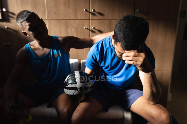 Dos jugadores de fútbol masculino de raza mixta que usan ropa deportiva sentados en el vestuario durante un descanso en el juego, sosteniendo la pelota descansando decepcionados. - foto de stock