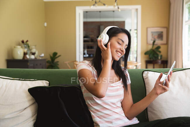 Mujer de raza mixta pasando tiempo en casa, usando auriculares y sosteniendo el teléfono inteligente en la sala de estar. Autoaislamiento y distanciamiento social en el bloqueo de cuarentena durante la epidemia de coronavirus covid 19. - foto de stock