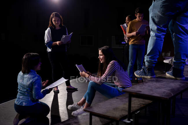 Vista lateral de un grupo multiétnico de adolescentes que tienen guiones y actúan en el escenario de un teatro escolar durante los ensayos para una actuación - foto de stock