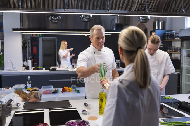 Caucasian female chef passing a leek to Caucasian male chef, with other chefs cooking in the background. Cookery class at a restaurant kitchen. — Stock Photo