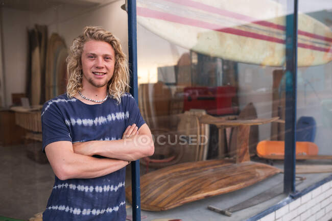 Creatore di tavole da surf maschio caucasico in piedi di fronte al suo studio, appoggiato su un telaio della porta dell'ingresso con le mani incrociate, guardando la fotocamera e sorridendo. — Foto stock
