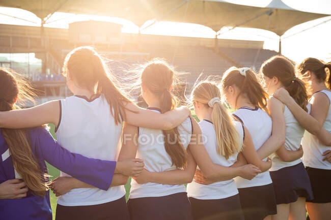 Rückansicht einer Gruppe kaukasischer Hockeyspielerinnen, die sich vor einem Spiel vorbereiten, an einem sonnigen Tag in einer Reihe stehend, die Hände auf den Schultern ihrer Teamkolleginnen umarmend — Stockfoto