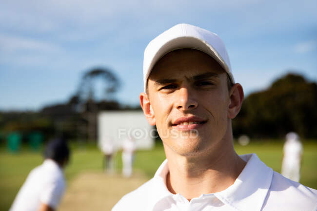 Портрет уверенного подростка, белого игрока в крикет, в белом и кепке, стоящего на поле для крикета в солнечный день, смотрящего в камеру, с другими игроками на заднем плане. — стоковое фото
