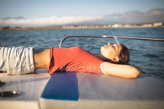 Una adolescente caucásica disfrutando de su tiempo de vacaciones en el sol junto a la costa, tumbada en un barco, relajándose, mirando hacia otro lado - foto de stock