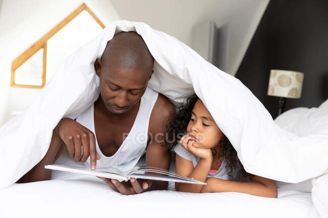 Front view of an African American man and his young daughter relaxing in the bedroom, lying under the covers and reading a book together — Stock Photo