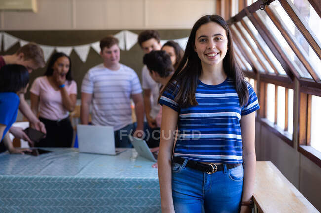 Porträt eines kaukasischen Teenagers mit langen, dunklen Haaren und braunen Augen, der in einem Klassenzimmer steht und in die Kamera lächelt, während sich Mitschüler im Hintergrund unterhalten — Stockfoto
