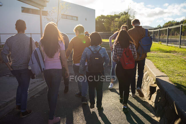 Rear view of a multi-ethnic group of male and female teenage students talking as they walk through their school grounds — Stock Photo