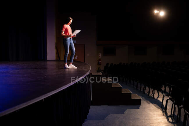 Vista laterale di un'adolescente caucasica in un teatro vuoto del liceo che si prepara prima di uno spettacolo, in piedi sul palco, con in mano una sceneggiatura e pratica la sua parte — Foto stock