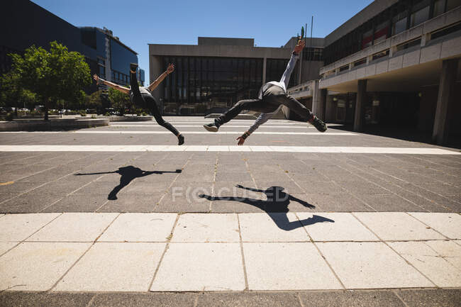 Vista traseira de dois homens caucasianos praticando parkour pelo edifício em uma cidade em um dia ensolarado, pulando acima do pavimento. — Fotografia de Stock