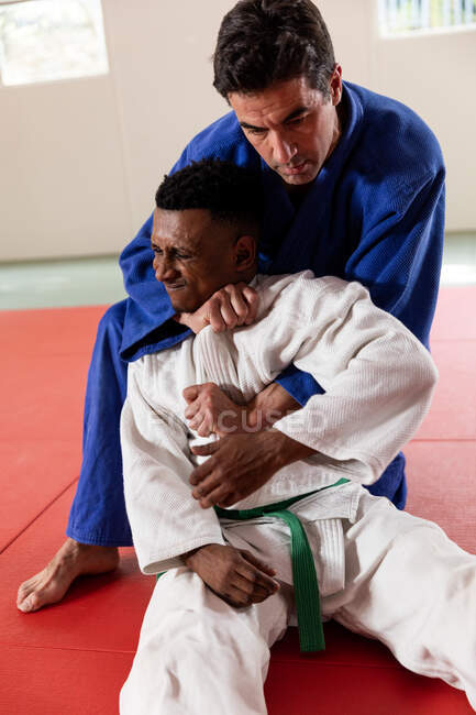 Vista frontale da vicino di un allenatore di judo maschile di razza mista e di un judoka maschile di razza mista adolescente con indosso judogi blu e bianchi, che pratica il judo durante un allenamento in palestra. — Foto stock