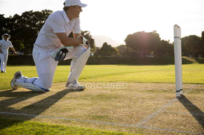 Vista laterale di un adolescente caucasico giocatore di cricket maschile con indosso bianchi e una tazza, inginocchiato vicino al wicket, sorridente e guardando la partita in campo durante una giornata di sole — Foto stock