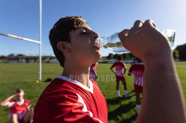 Seitliche Nahaufnahme eines jugendlichen kaukasischen männlichen Rugby-Spielers in rot-weißer Mannschaftskleidung, der auf einem Spielfeld steht und Wasser trinkt, mit den anderen Spielern im Hintergrund. — Stockfoto