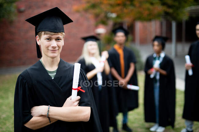 Portrait of teenage Caucasian male high school student wearing a cap and gown, holding a diploma on his graduation day, looking to camera and smiling, with other students wearing caps and gowns in the background — Stock Photo