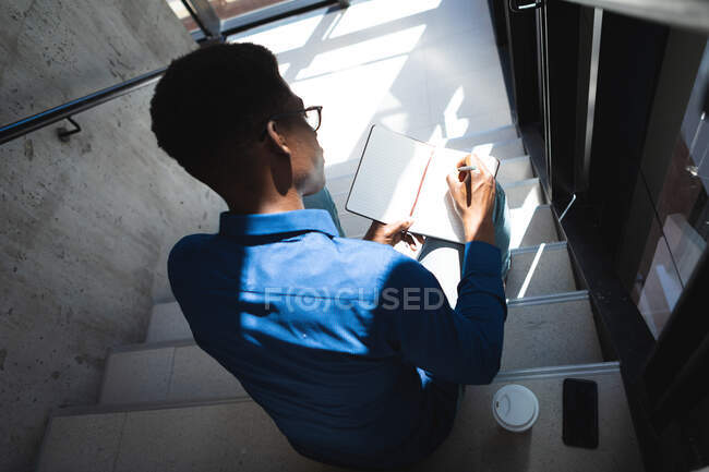 Афроамериканский бизнесмен с темными короткими волосами, в синей рубашке и очках, работает в современном офисе, сидит на лестнице и пишет заметки — стоковое фото