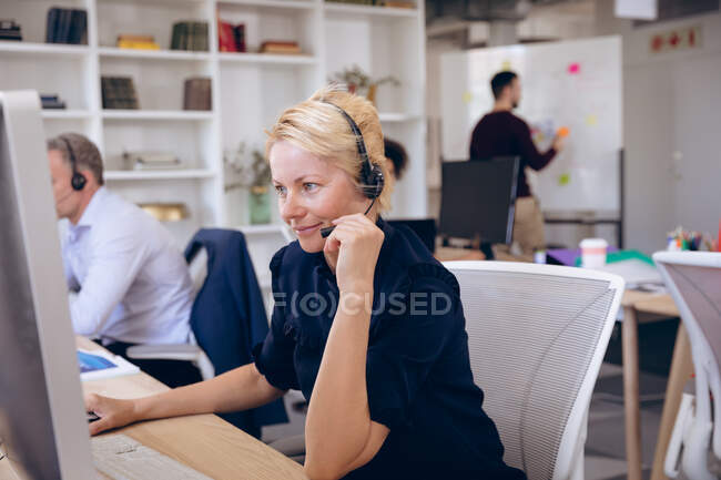 Una mujer de negocios caucásica trabajando en una oficina moderna, sentada en un escritorio, usando una computadora portátil, usando auriculares y hablando por teléfono, con sus colegas de negocios trabajando en segundo plano - foto de stock