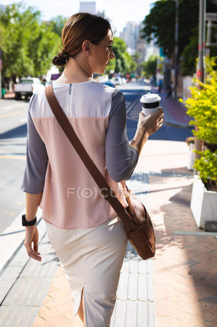Uma mulher de negócios caucasiana em movimento em um dia ensolarado, andando em uma rua da cidade e segurando um café takeaway — Fotografia de Stock