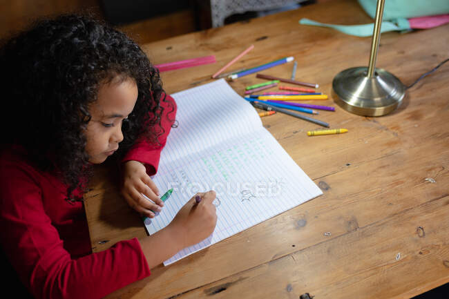 Blick aus der Vogelperspektive auf ein junges afroamerikanisches Mädchen zu Hause, das am Esstisch sitzt und ihre Hausaufgaben macht und in ein Schulbuch schreibt — Stockfoto