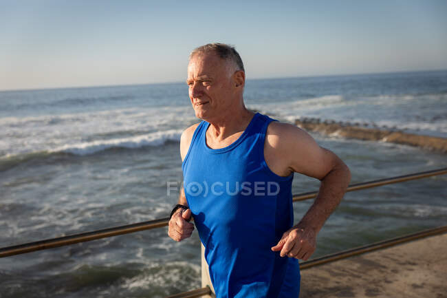 Vista lateral de un hombre mayor caucásico maduro haciendo ejercicio en un paseo marítimo en un día soleado con cielo azul, corriendo - foto de stock