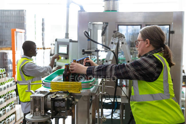 Caucasian man wearing high visibility vest, working in a microbrewery, checking bottles of beer with an African American man working in the background. — Stock Photo