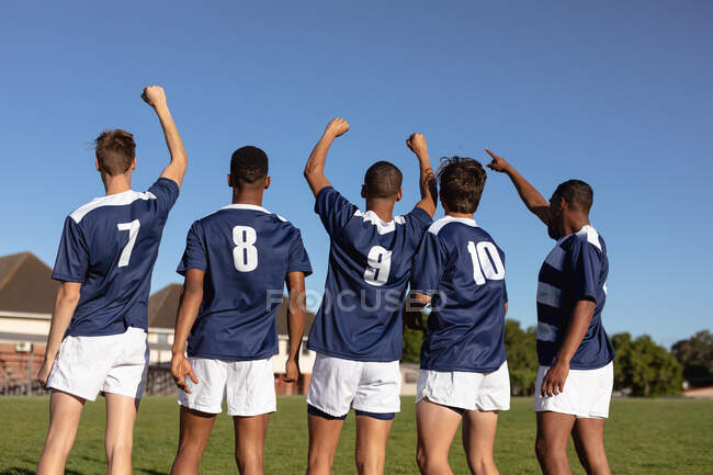 Rückansicht einer Gruppe multiethnischer männlicher Rugby-Spieler mit blau-weißer Mannschaftskleidung, die auf einem Spielfeld stehen, die Hände heben und während eines Spiels jubeln — Stockfoto