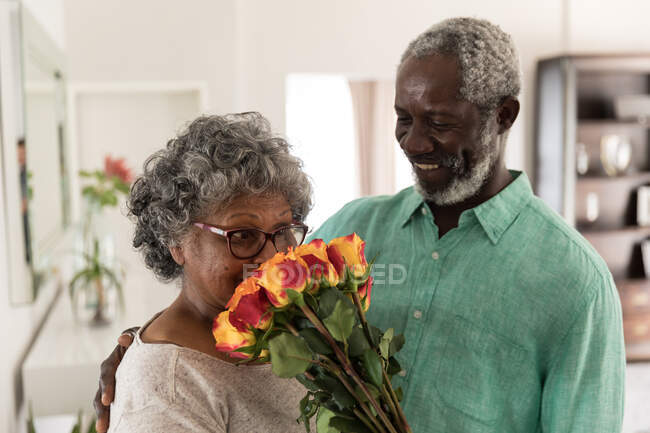 Ein älteres afroamerikanisches Paar, das während der Coronavirus-Epidemie 19 Zeit zu Hause verbringt, soziale Distanzierung und Selbstisolierung in Quarantäne, der Mann hält einen Blumenstrauß in der Hand, lächelt und schenkt ihn der Frau — Stockfoto