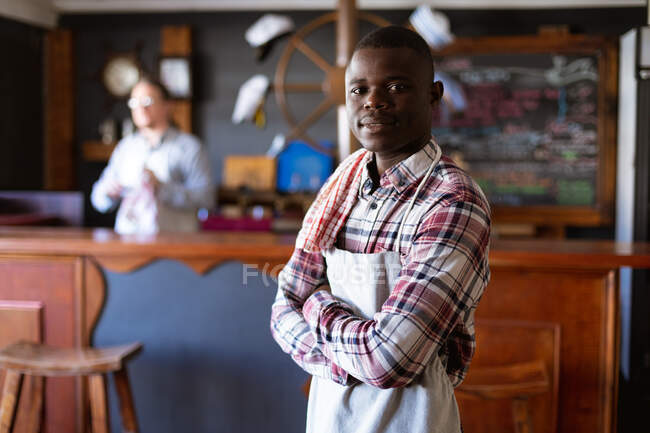 Ritratto di un barista afroamericano con grembiule bianco, che lavora in un pub di microbirreria, con le braccia incrociate e guarda dritto in una macchina fotografica. — Foto stock