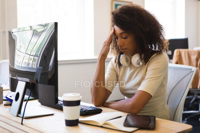 Una mujer de negocios de raza mixta con el pelo rizado, trabajando en una oficina moderna, sentada en una mesa y tocándose la frente - foto de stock