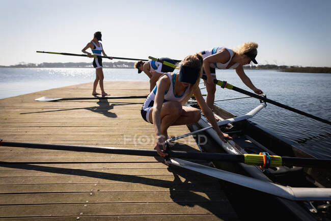 Vista laterale di una squadra di canottaggio di quattro donne caucasiche che si allenano sul fiume, su un molo al sole che preparano una barca prima di remare — Foto stock