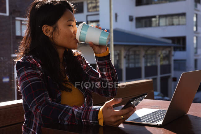 Vue latérale d'une femme métisse aux longs cheveux foncés assise à une table dans un café pendant la journée, travaillant sur un ordinateur portable à l'aide d'un smartphone buvant du café à emporter avec des bâtiments en arrière-plan. — Photo de stock