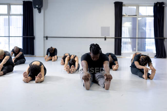 Vue latérale d'un groupe multi-ethnique de danseurs modernes en forme, hommes et femmes, portant des tenues noires pratiquant une routine de danse pendant un cours de danse dans un studio lumineux, assis sur le sol et s'étirant vers le haut. — Photo de stock
