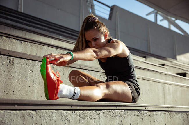 Vista lateral de uma atleta caucasiana praticando em um estádio de esportes, sentada nas arquibancadas e alongada, segurando o pé — Fotografia de Stock